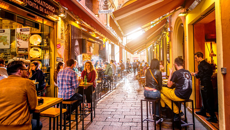 People sit on bar stools outside on brick alleyway eating lunch on Pescherie Vecchie street near famous Italian food markets and shops in the center of Bologna, Italy.