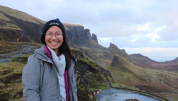 A student smiles for a photo in front of a grassy mountain range and a body of water behind them. 