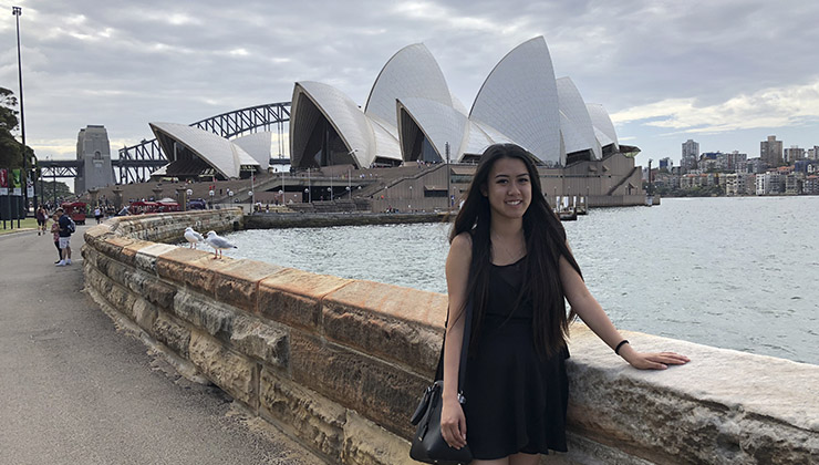 Student smiles for the camera with a view of the Sydney Opera House and Sydney Harbor in Australia. 