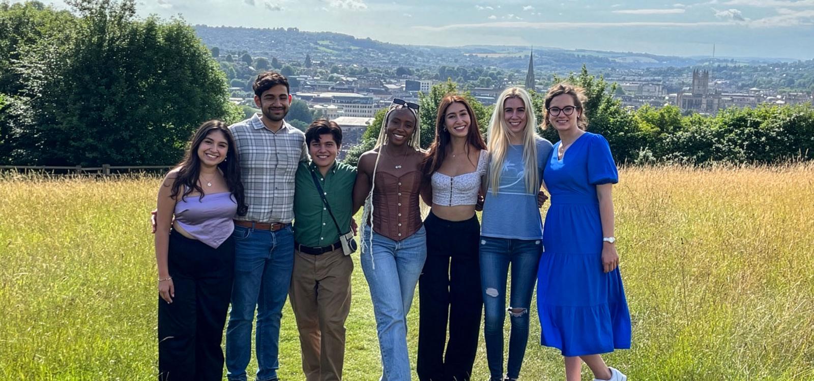 Seven students pose for a photo atop a grassy hill with historic English architecture behind them in the distance.