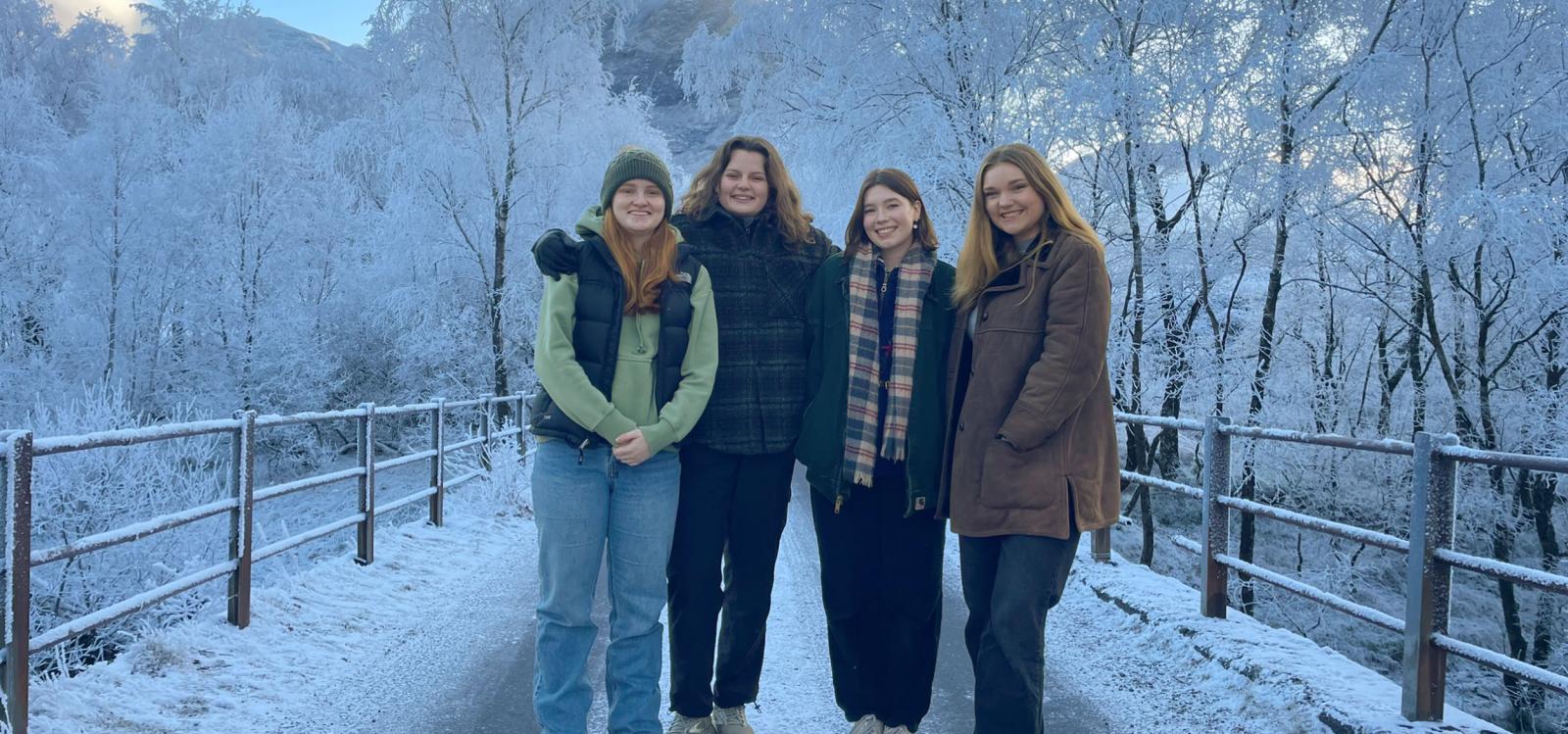 Four students dressed in coats, scarves, and boots stand on a snow-covered bridge.